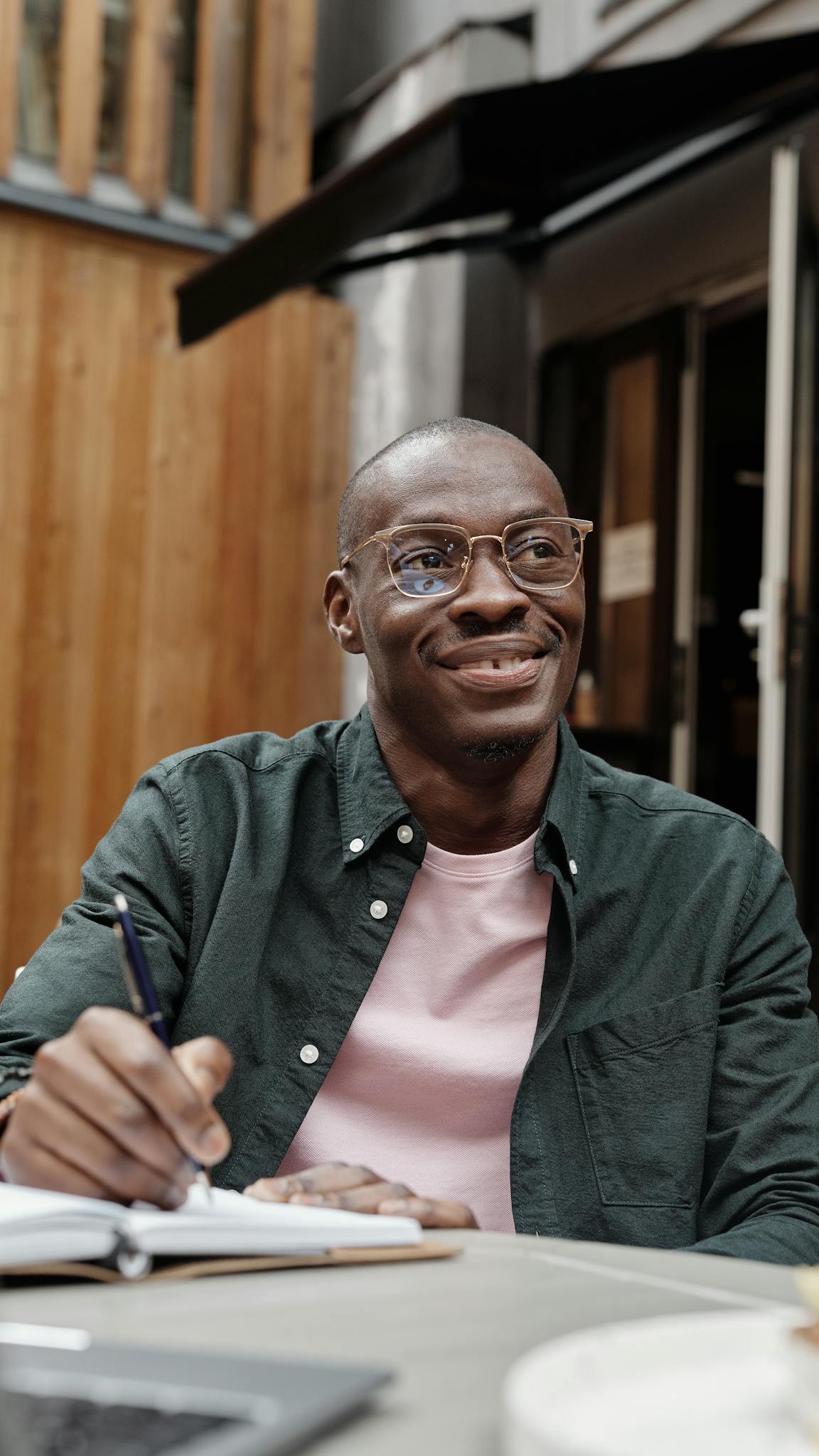 A smiling man wearing glasses, a dark green shirt, and a pink t-shirt writes in a notebook while sitting at an outdoor table. A building with wooden panels is visible in the background.