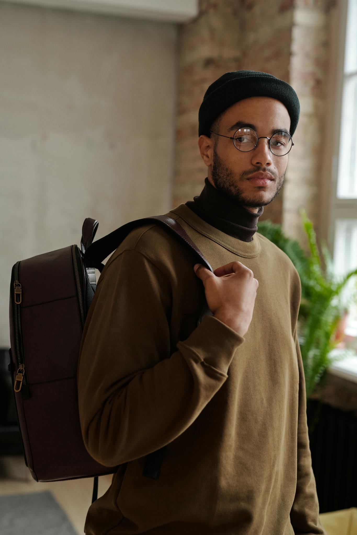 A man wearing glasses, a black beanie, and a brown sweatshirt stands indoors with a backpack over his shoulder, looking at the camera. There is a window and a green plant in the background.