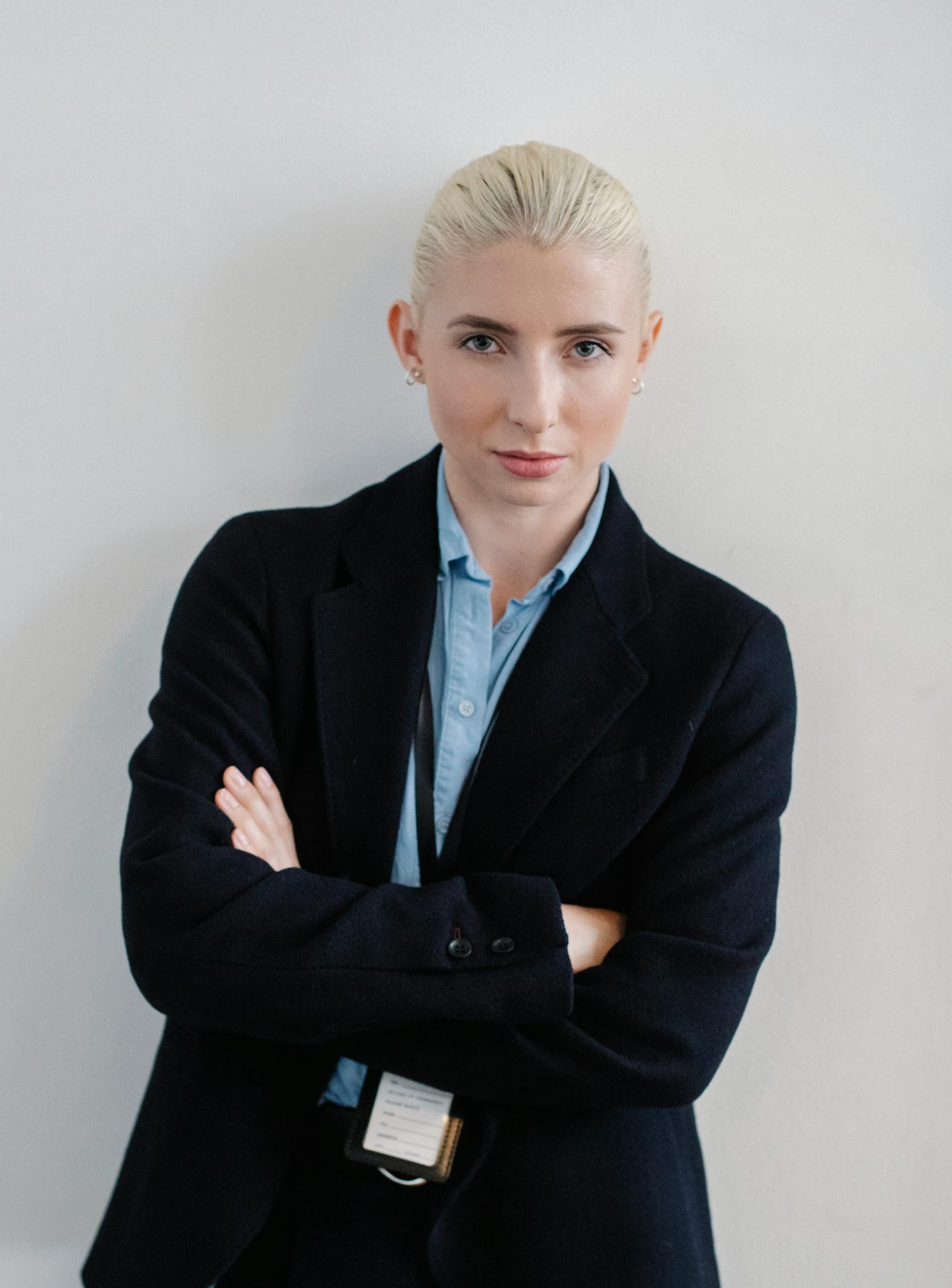 A person with blonde hair tied back, wearing a dark blazer and light blue shirt, stands against a white wall with arms crossed, looking confidently at the camera.