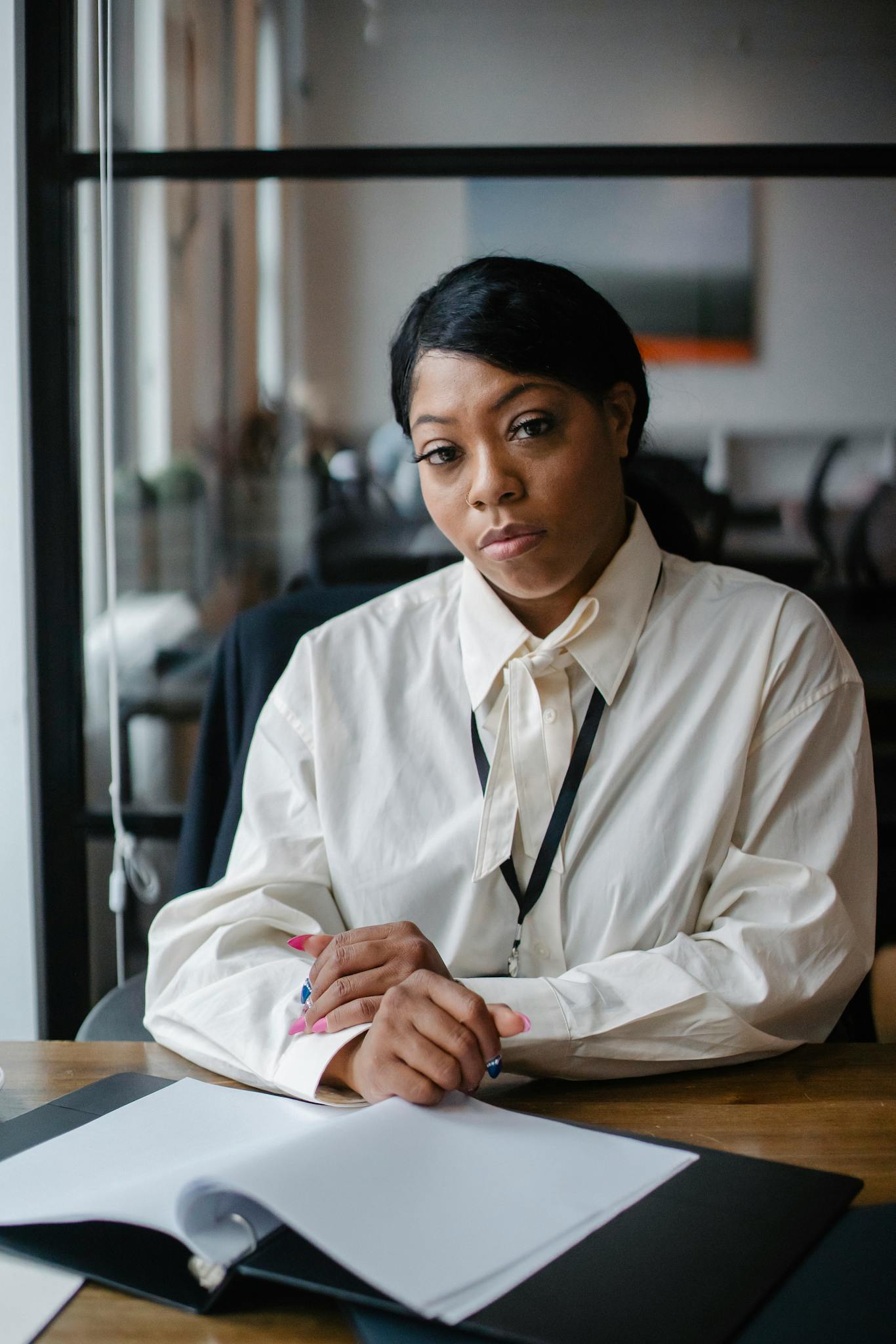 A woman wearing a white blouse and lanyard sits at a desk with her hands on an open notebook, looking thoughtfully at the camera in a modern office setting.